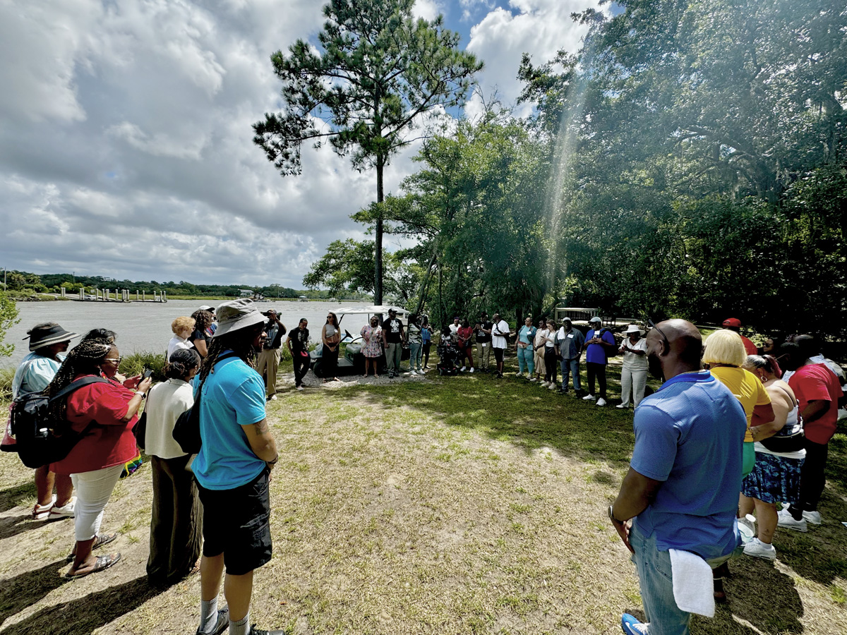 Dozens of people standing in circle outside near trees and water.