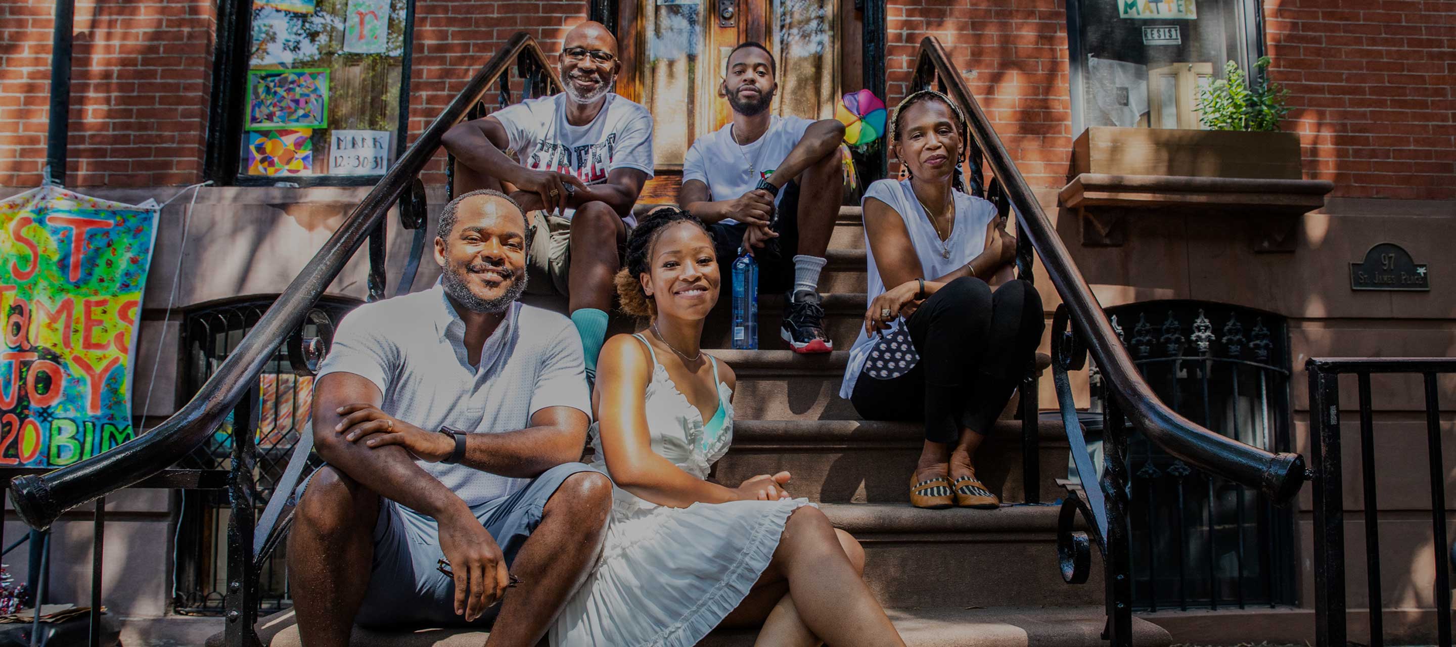 Photo of three Black men and two Black women sitting on the steps of a brownstone house.